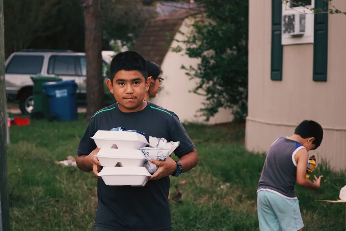 A boy carrying a stack of hot meal boxes across a yard, another child behind him with a drink.