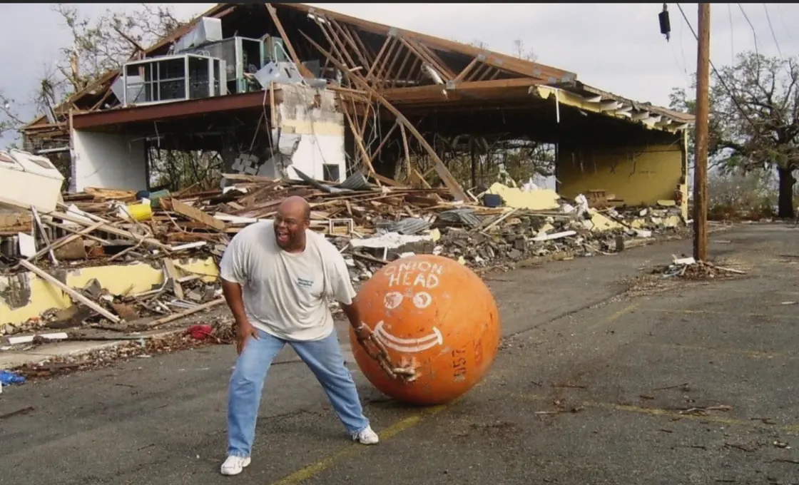 A man laughing as he rolls a giant orange buoy painted with a smiley face down a street, past a building flattened to its rafters.