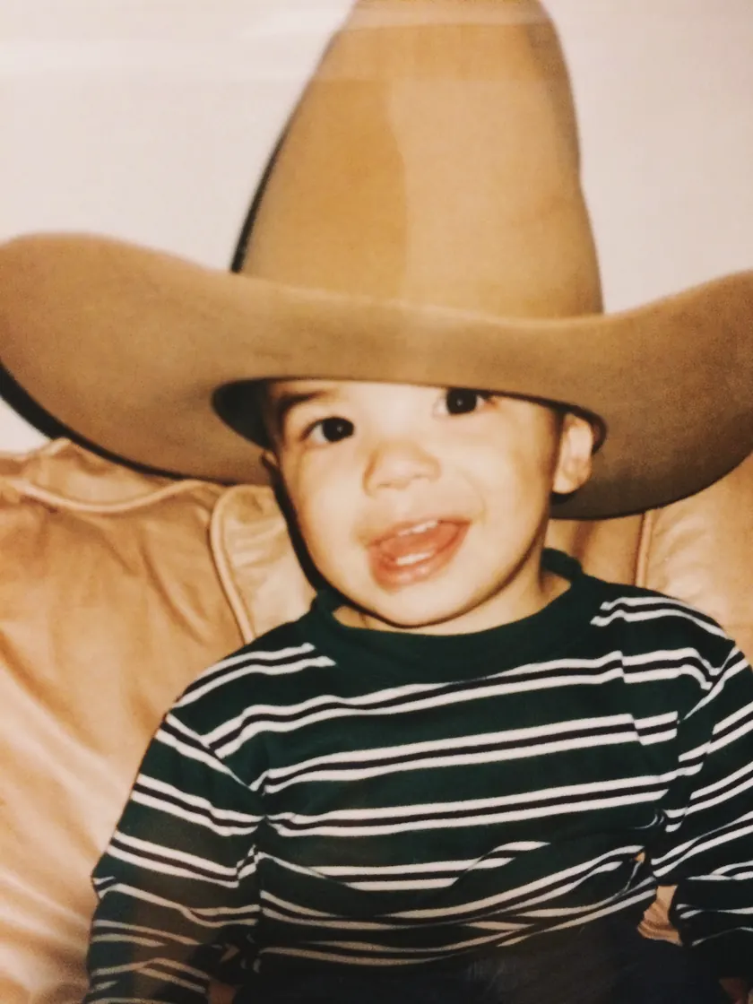 Me as a toddler in a striped shirt, grinning under a cowboy hat far too big for me.