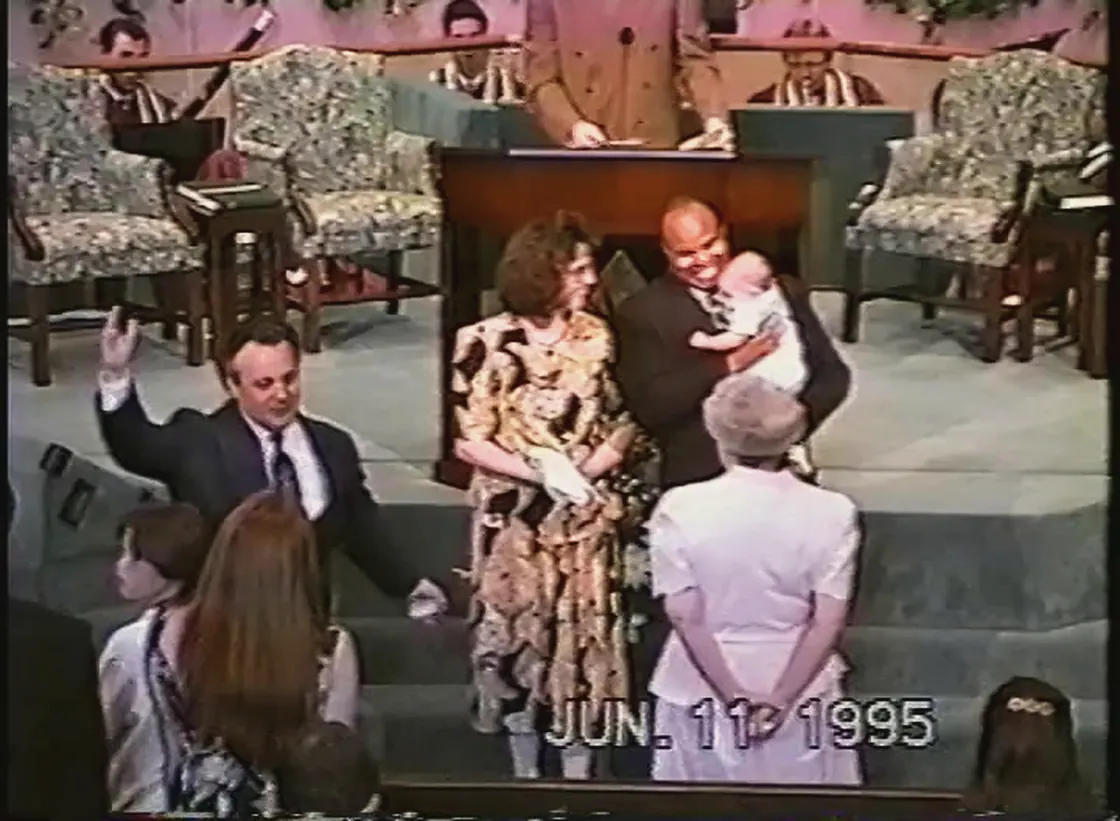 A man in a dark suit holding a baby in white at the front of a church, a woman in a patterned dress beside him, and a man in a suit to the left reading from something in his hands.
